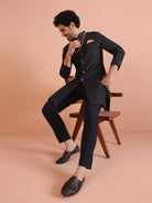 The man is seated gracefully on a chair, posing in a navy blue embellished Indo-Western set