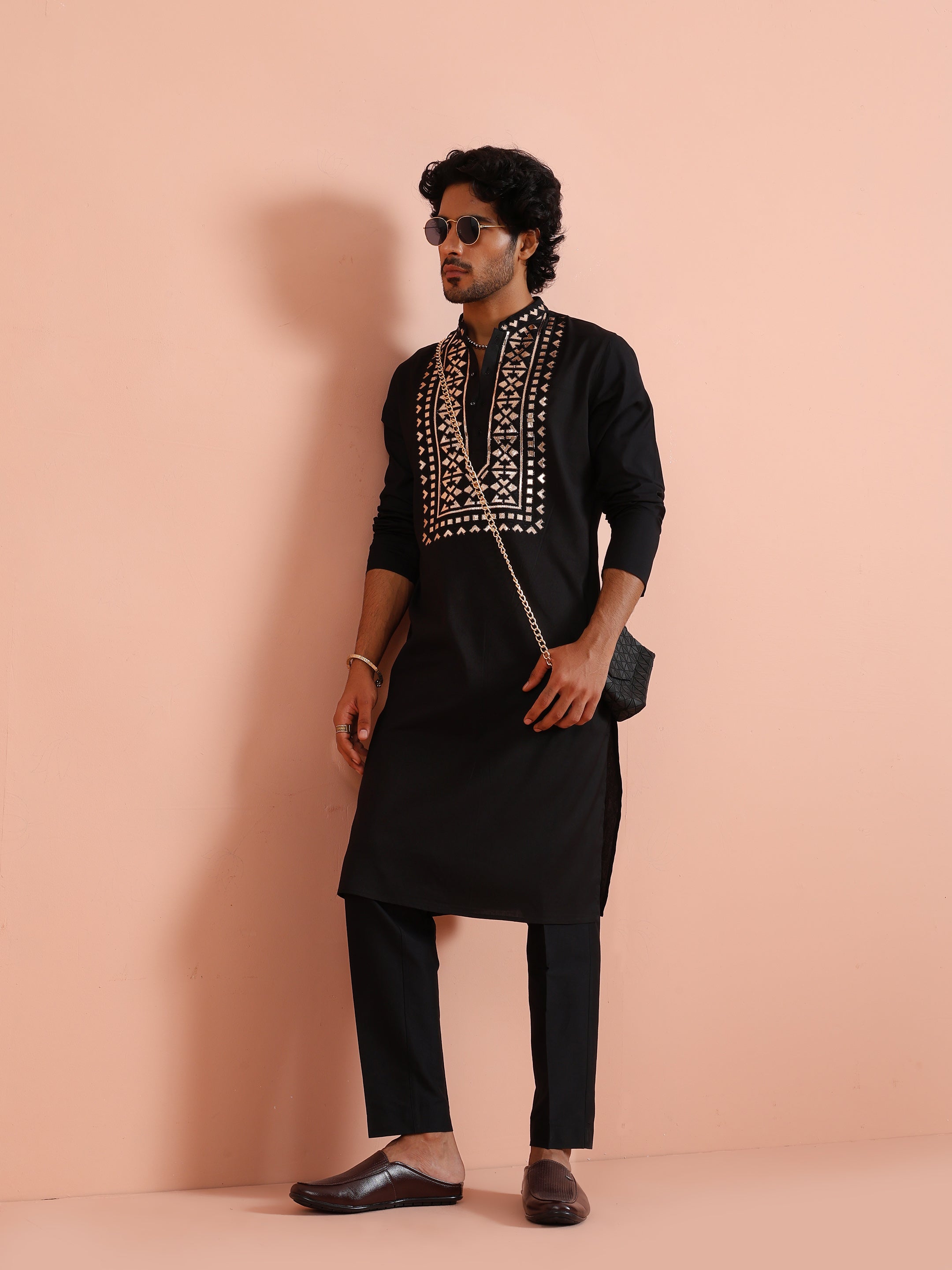 Dressed in a Black Metallic Embroidered Kurta Set, the man strikes a stylish side pose with sunglasses and a crossbag
