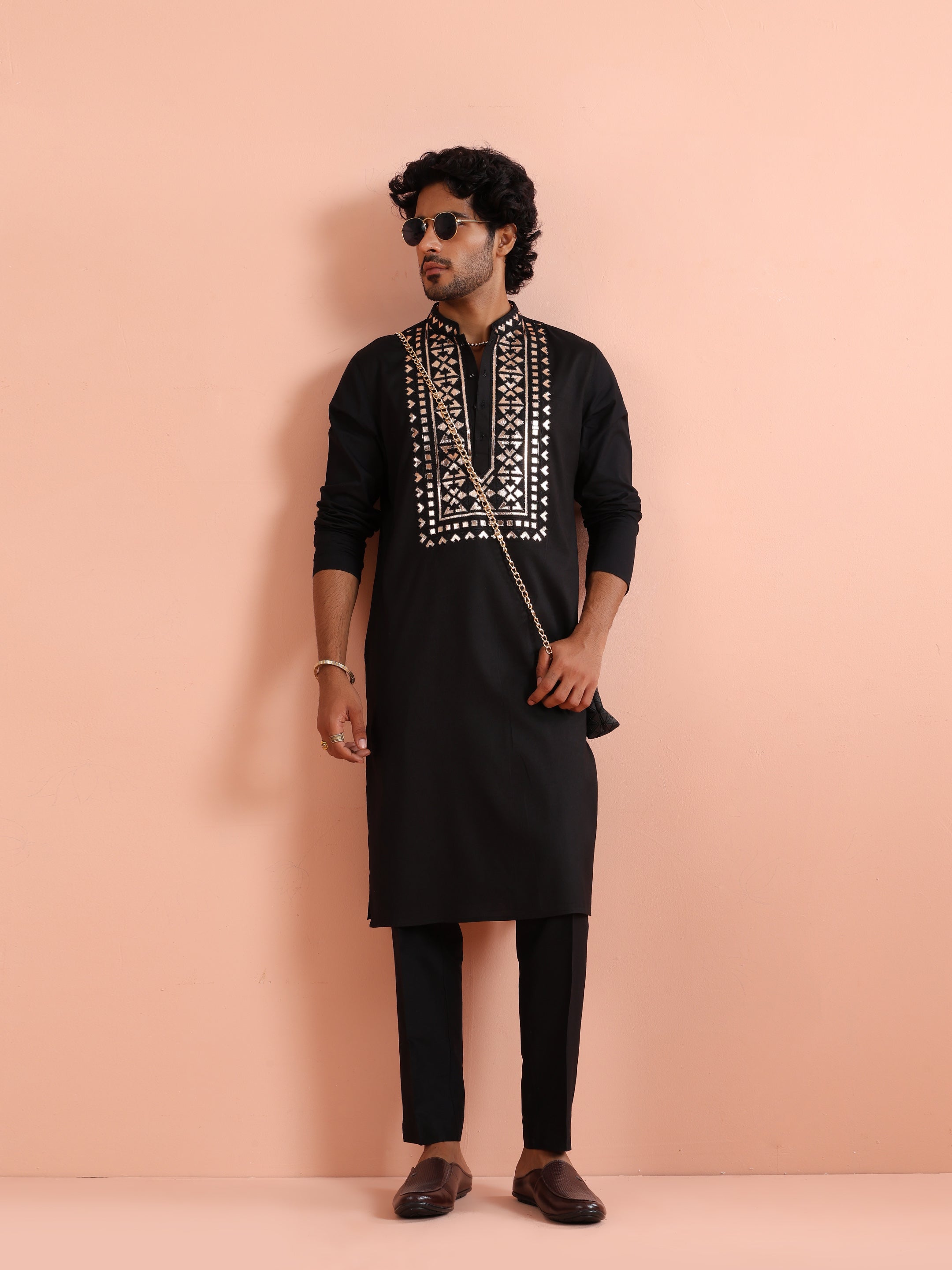 Dressed in the Black Metallic Embroidered Kurta Set, the model looks to his right with sunglasses and a crossbag